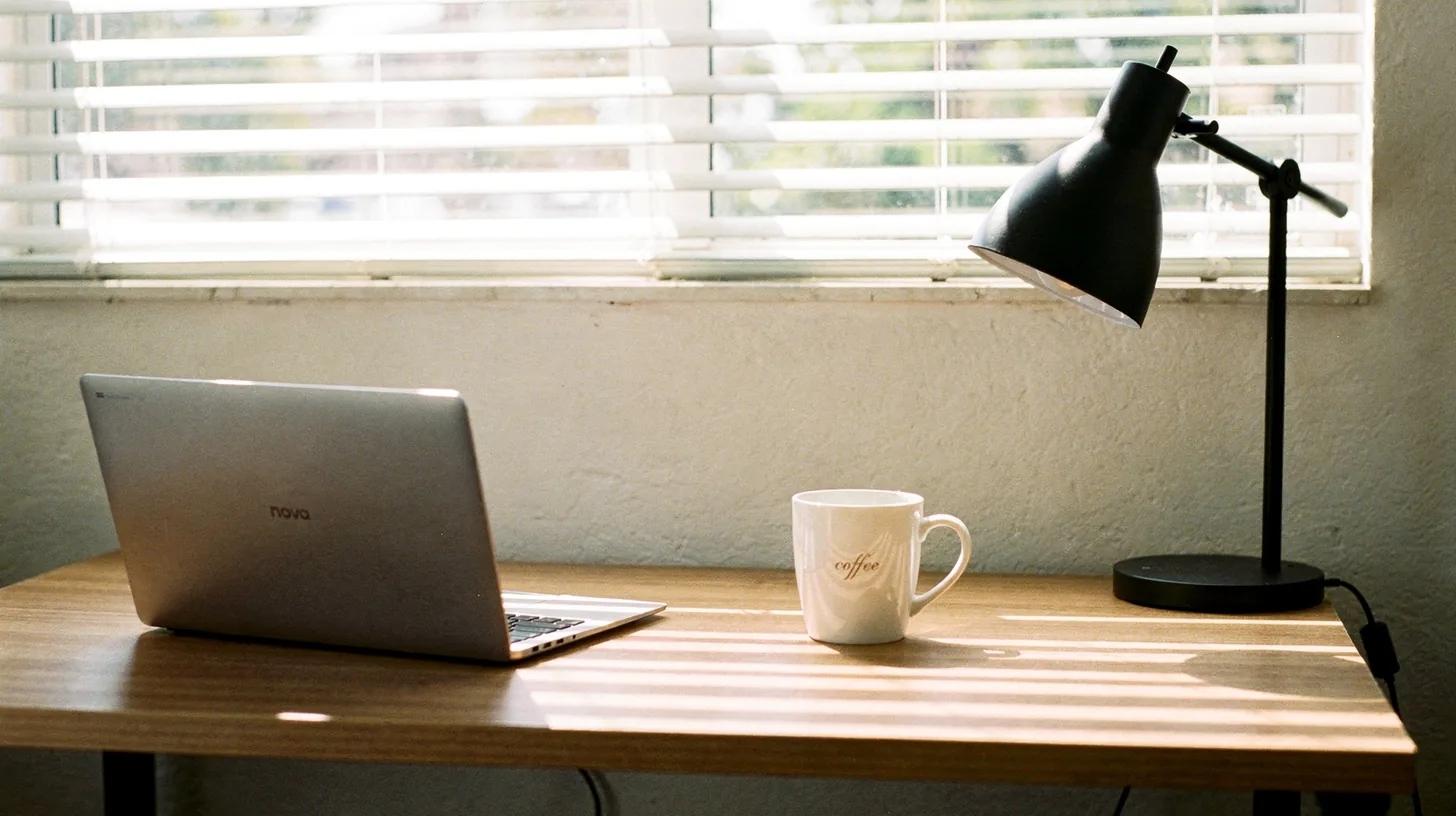An empty small-office desk by a window with a closed laptop and a mug, venetian blinds throwing hard bars of light across the surface